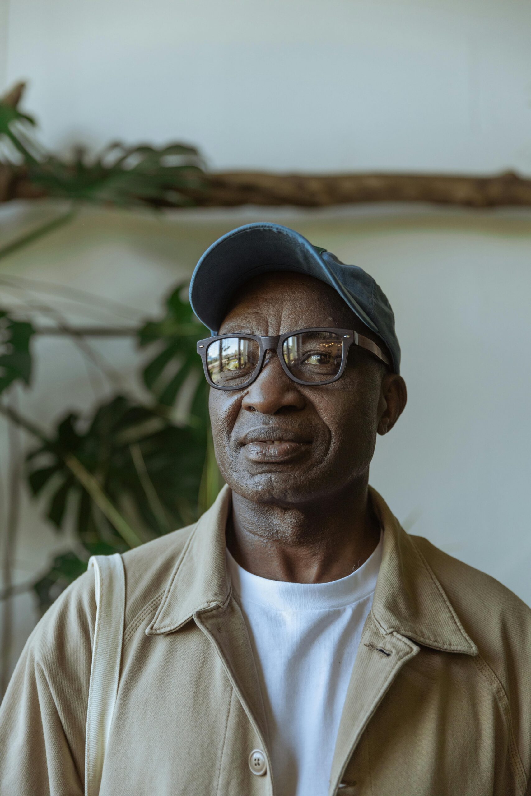 Senior man smiling indoors wearing glasses and a baseball cap with indoor plants in the background.